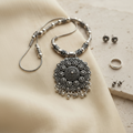 An ornate silver-toned necklace with a large circular floral pendant and matching beaded chain, styled on a beige fabric surface with small jewelry props nearby.